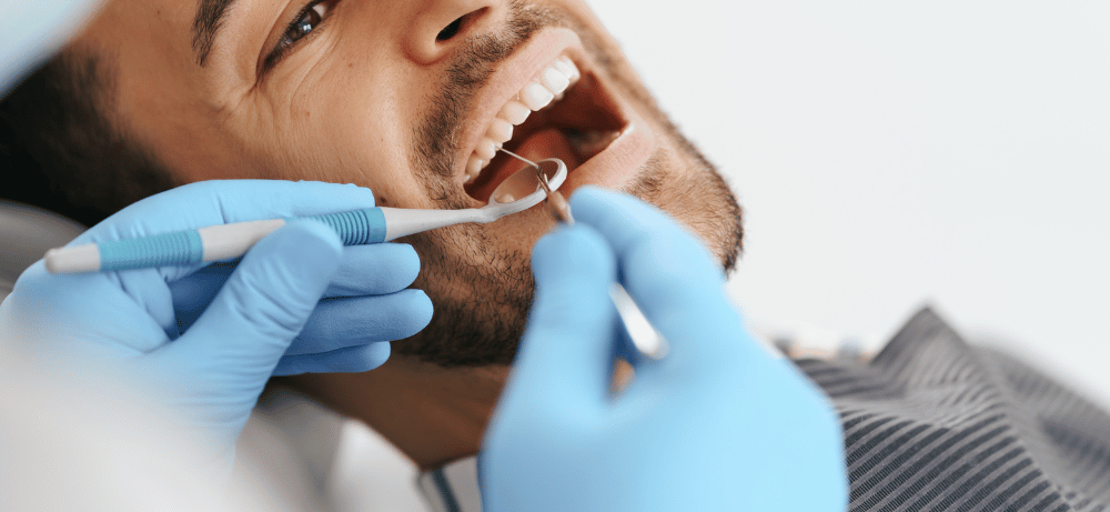 Man ligger i en tandläkarstol och får sina tänder undersökta av en tandläkare i blå handskar med tandinstrument.